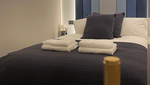 A neatly made bed with folded towels, a bedside table with a lamp, and a modern headboard with a blue and white color scheme.
