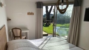 Une chambre avec un grand lit, une petite table et une chaise. Une porte-fenêtre coulissante donne sur un jardin.