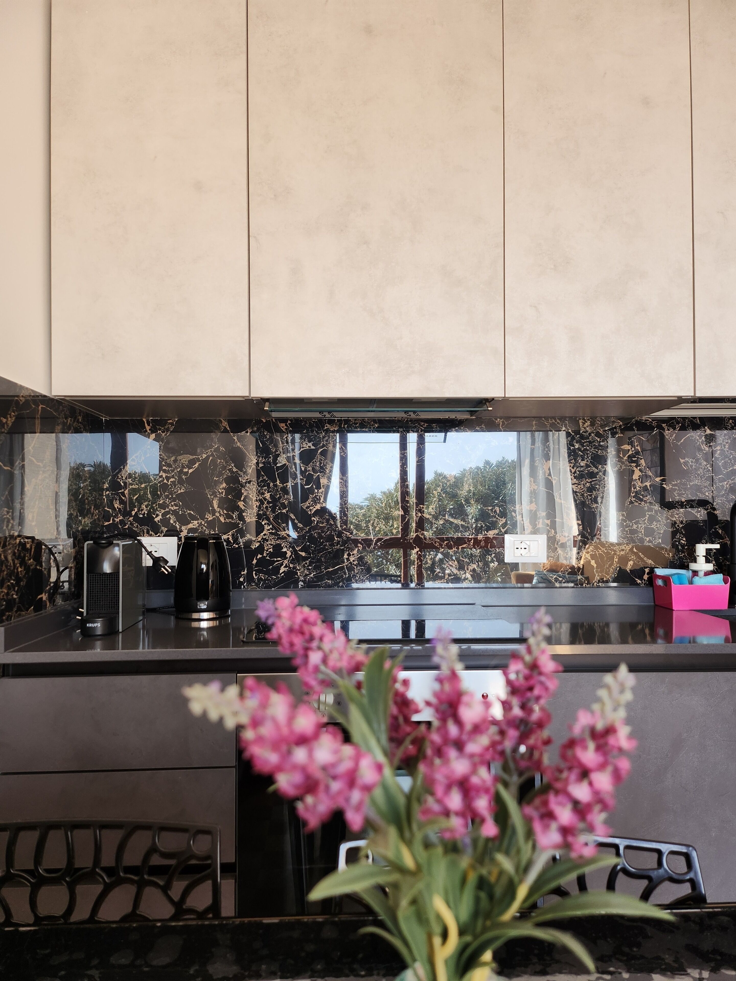 Een moderne keuken met een marmeren spatwand, een koffiezetapparaat en een bloemstuk met roze bloemen.