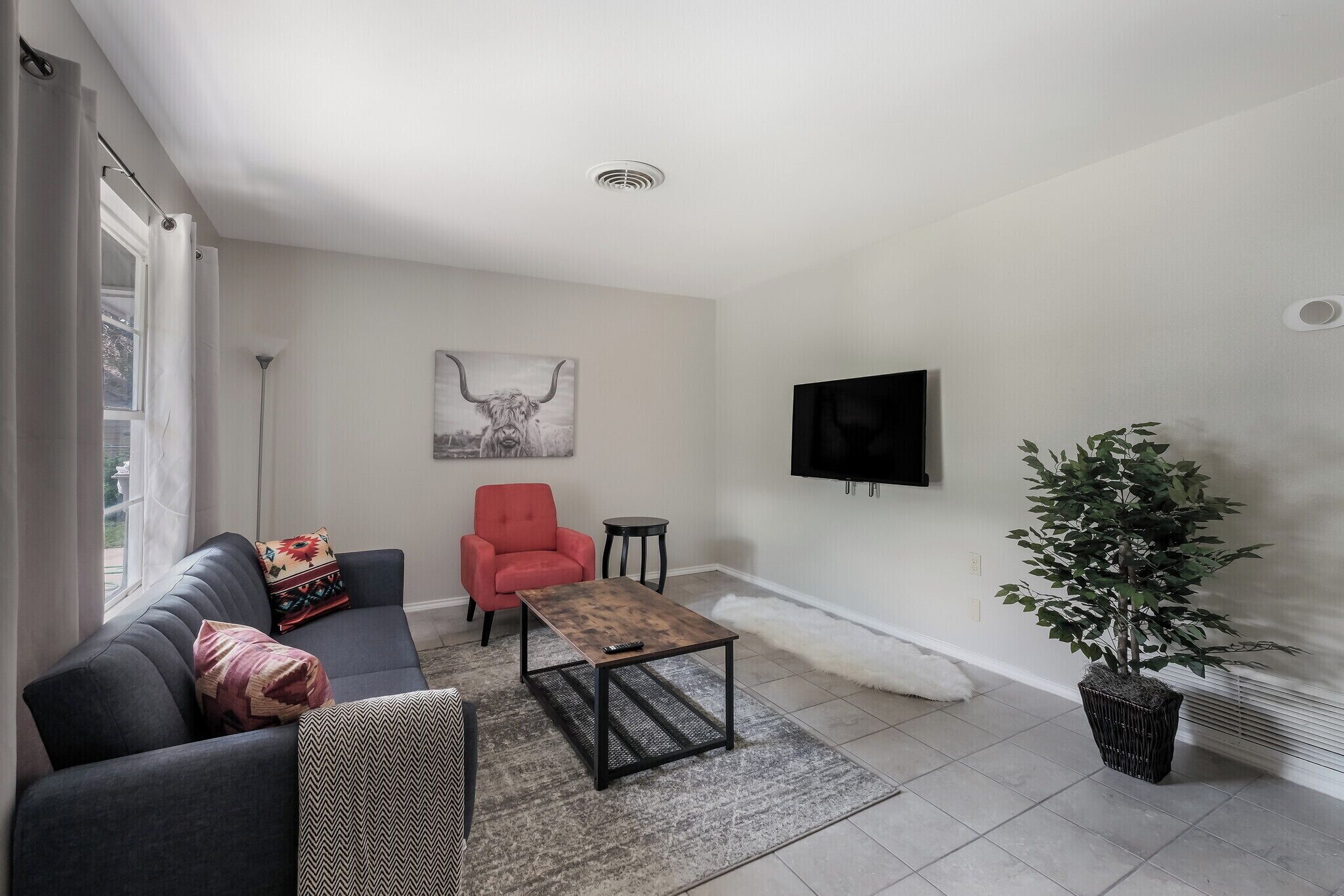 Uma sala de estar com um sofá cinzento, uma poltrona vermelha, uma mesa de centro, uma TV fixada na parede e um vaso com planta.