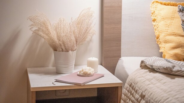 A neatly made bedside table with a white vase containing dried pampas grass, a white candle, and a pink notebook.