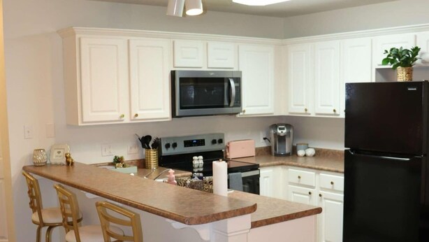 A kitchen with white cabinets, a black refrigerator, a microwave, a stove, and a sink. There are three stools at the island. - The Grace at the Highlands - Charming 2-bedroom townhome close to MSU campus (Starkville)