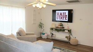 A living room with a ceiling fan, a mounted TV displaying "HAIR STATE", a sofa, a coffee table, and a small plant on a console table. - The Grace at the Highlands - Charming 2-bedroom townhome close to MSU campus (Starkville)