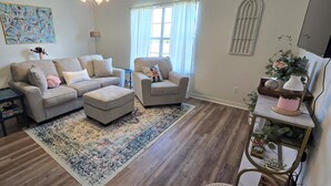 A living room with a sofa, armchair, and ottoman, a rug, a small table with a lamp, and a plant on a stand. - The Grace at the Highlands - Charming 2-bedroom townhome close to MSU campus (Starkville)