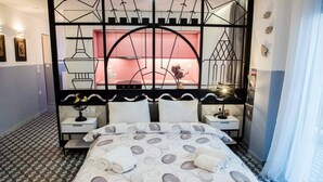 Egy hotelszoba geometriai mintázatú padlóval, fehér és szürke színű ágyneművel, valamint fekete fém ágykerettel, melynek a háttérfalát egy élénk rózsaszín színnel emelték ki.