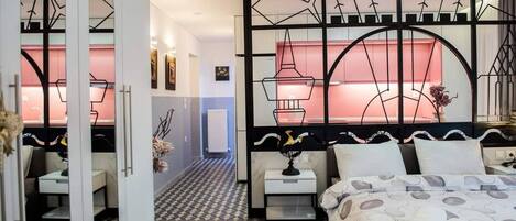 Habitación de hotel con piso de diseño geométrico, cama con cubrecama estampado, mesita de noche con un globo terráqueo y una ventana grande con cortinas rojas.