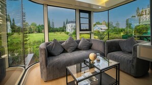 A modern living room with a large gray sofa, a glass coffee table, and a view of a lush garden and neighboring houses. - PARACH RESORT KHAOKHO (Khao Kho)