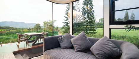 A modern living room with a gray sofa, two cushions, a glass coffee table, and a view of a lush green landscape through large windows.