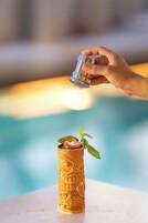 Una mano sirve una bebida en un vaso dorado de diseño intrincado, adornado con una hoja de menta, sobre una superficie blanca cerca de una piscina.