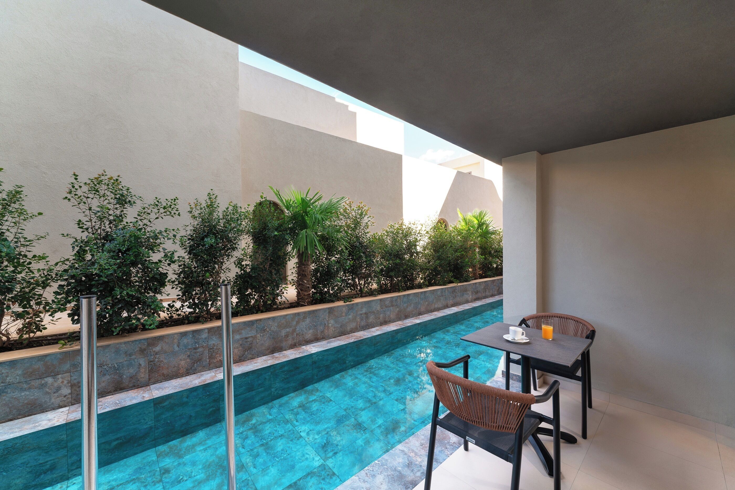 A poolside area with a table and chairs, surrounded by greenery and a clear pool.