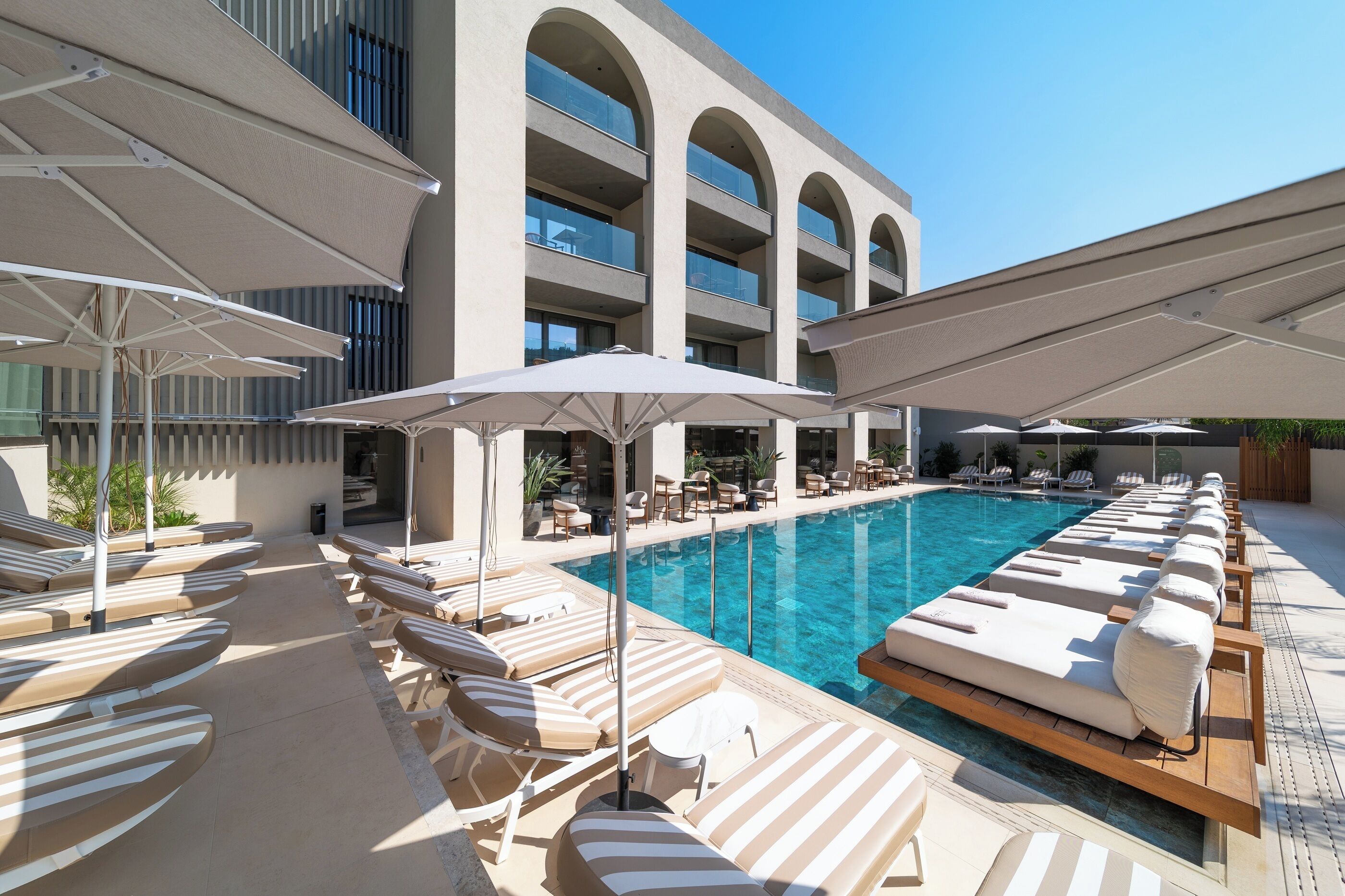 Un área junto a la piscina con sillones de descanso y sombrillas, una piscina azul cristalina y un edificio con varios balcones.