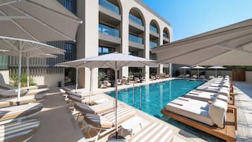 Un área junto a la piscina con sillones de descanso y sombrillas, una piscina azul cristalina y un edificio con varios balcones.