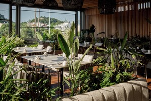 Un espace extérieur aménagé pour les repas, avec des tables et des chaises, entouré de grandes plantes vertes.