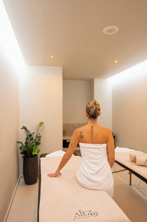 Una persona sentada en una camilla de masaje en un cuarto de spa, con una planta y una toalla a un lado.