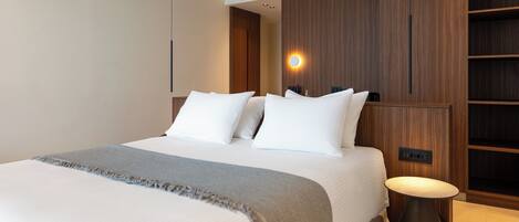 A modern hotel room with a large bed, a bedside table, and built-in wooden shelving.