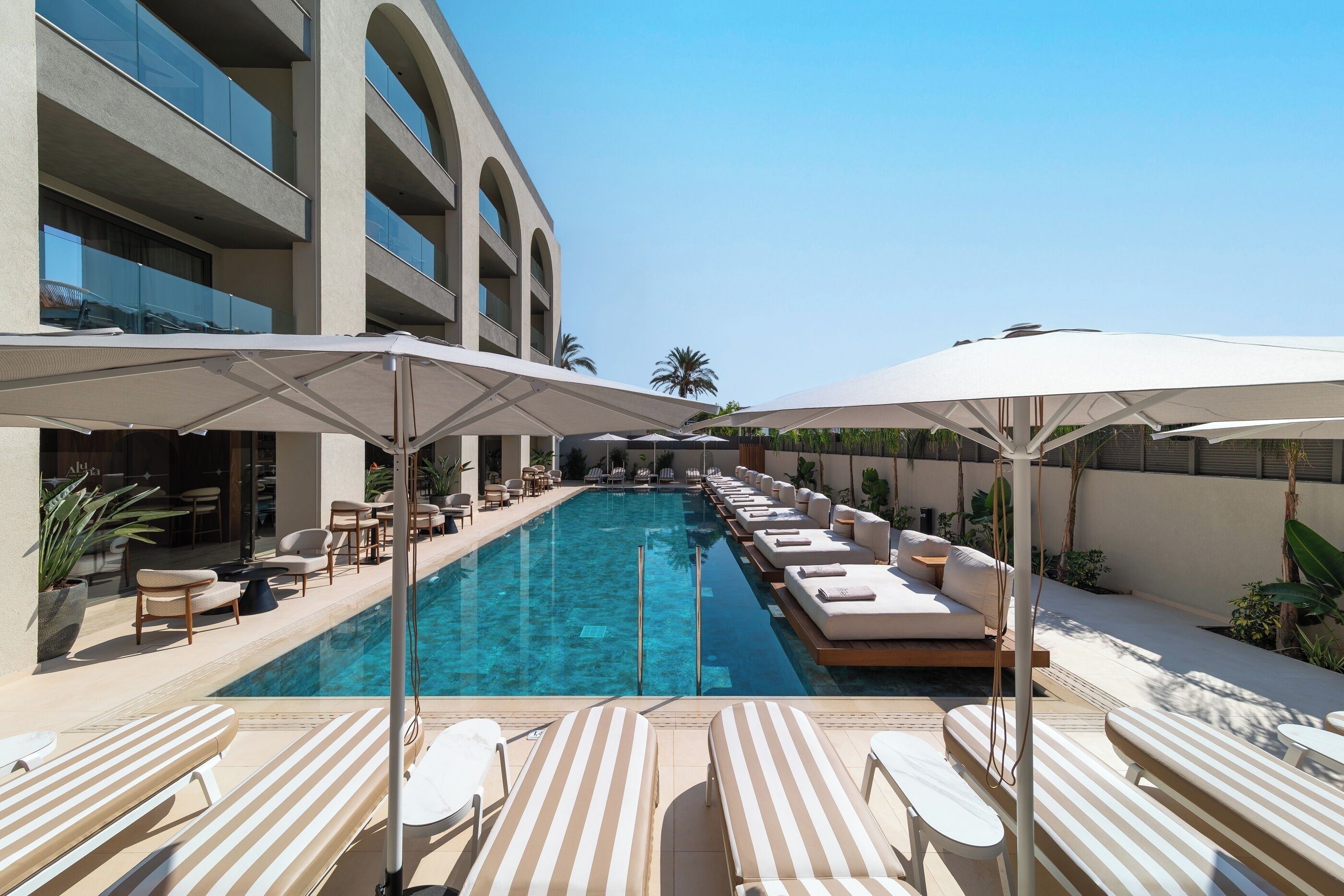 Un área de piscina con sillones reclinables y sombrillas, rodeada por un edificio de varios pisos con balcones.
