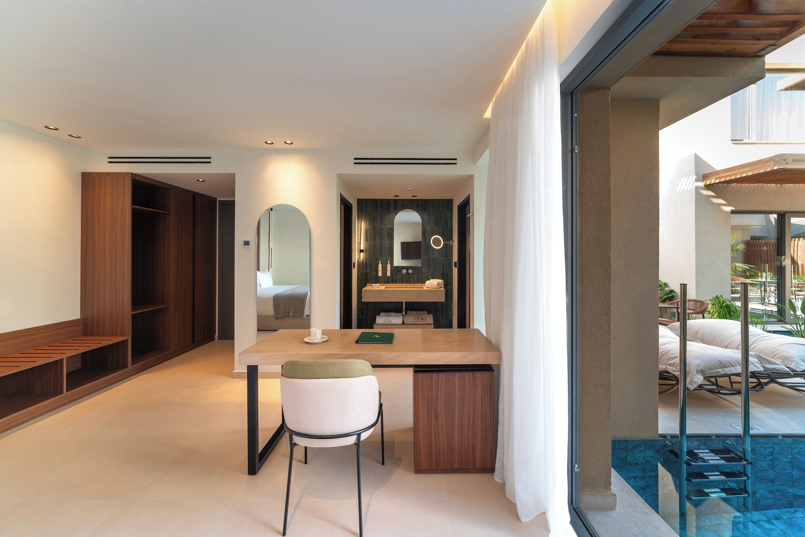 A modern hotel room with a dining area, a large mirror, a bathroom with a sink and mirror, and a view of the outdoor pool area.
