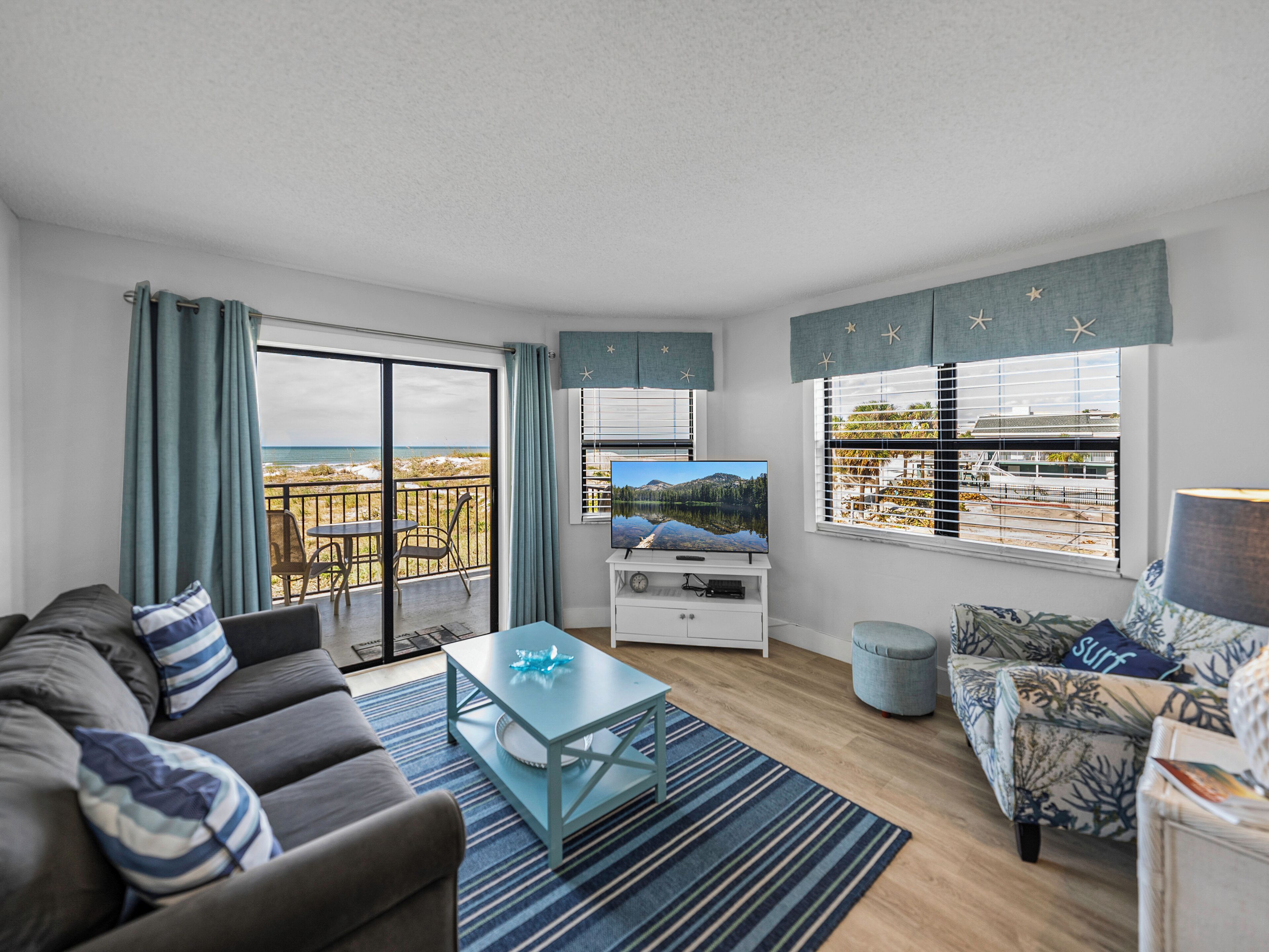 A modern living room with a sofa, a coffee table, and a TV on a white cabinet. There is a balcony with a view of the beach and a dining area with chairs.