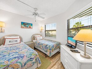 A hotel room with two beds, a ceiling fan, a painting, a window with blinds, and a desk with a lamp.