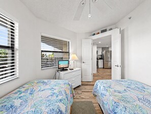 Ein Schlafzimmer mit zwei Betten, einem Schreibtisch mit Computer, einem Fenster mit Jalousien und Blick ins Freie.