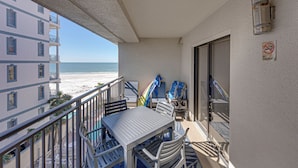 Een balkon met een tafel en stoelen, uitkijkend op een strand en een gebouw met veel ramen.