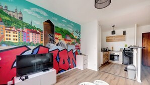 A modern apartment with a vibrant cityscape mural, a flat-screen TV, a dining area with white stools, and a kitchen with wooden cabinets and a black countertop. - Au Lyon D'ort - Near Eurexpo (Villeurbanne)