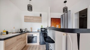 A modern kitchen with wooden cabinets, a built-in oven, a microwave, and a black bar stool. - Au Lyon D'ort - Near Eurexpo (Villeurbanne)