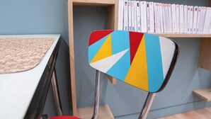 Une chaise ornée d’un motif géométrique coloré, une étagère en bois remplie de livres et une table sur laquelle est posée une nappe tressée.