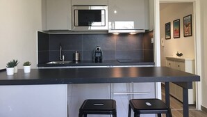 A modern kitchen with grey cabinets, a black countertop, a built-in microwave, a coffee machine, and two black stools. - To complete (Lyon)