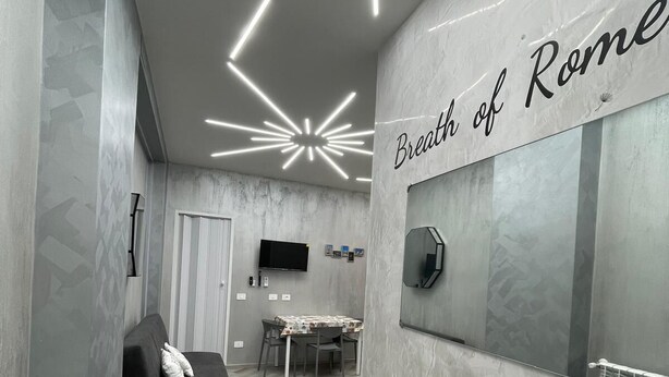 A modern room with a black sofa, a dining table, and a large mirror with the text "Breath of Rome" on the wall.