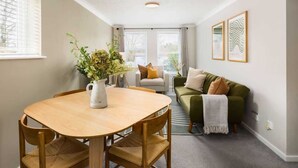 Un salon moderne avec une table à manger en bois, un canapé vert et une table basse. Deux œuvres d’art encadrées ornent le mur, et une fenêtre est dotée de stores.