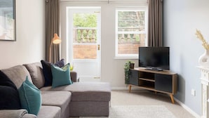 A modern living room with a grey sectional sofa, a flat-screen TV on a wooden stand, and a large window with curtains. - Modern | 2 Bed | Chestnut Court Leamington Spa (Royal Leamington Spa)