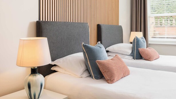 A hotel room with two beds, a nightstand with a lamp, and a window with curtains. - Modern | 2 Bed | Chestnut Court Leamington Spa (Royal Leamington Spa)