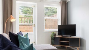 A modern living room with a grey sofa, a flat-screen TV on a wooden stand, and a window with curtains. - Modern | 2 Bed | Chestnut Court Leamington Spa (Royal Leamington Spa)