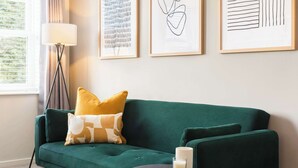 Un sofá verde con un cojín amarillo, una mesita auxiliar negra con una vela, y cuadros en la pared.