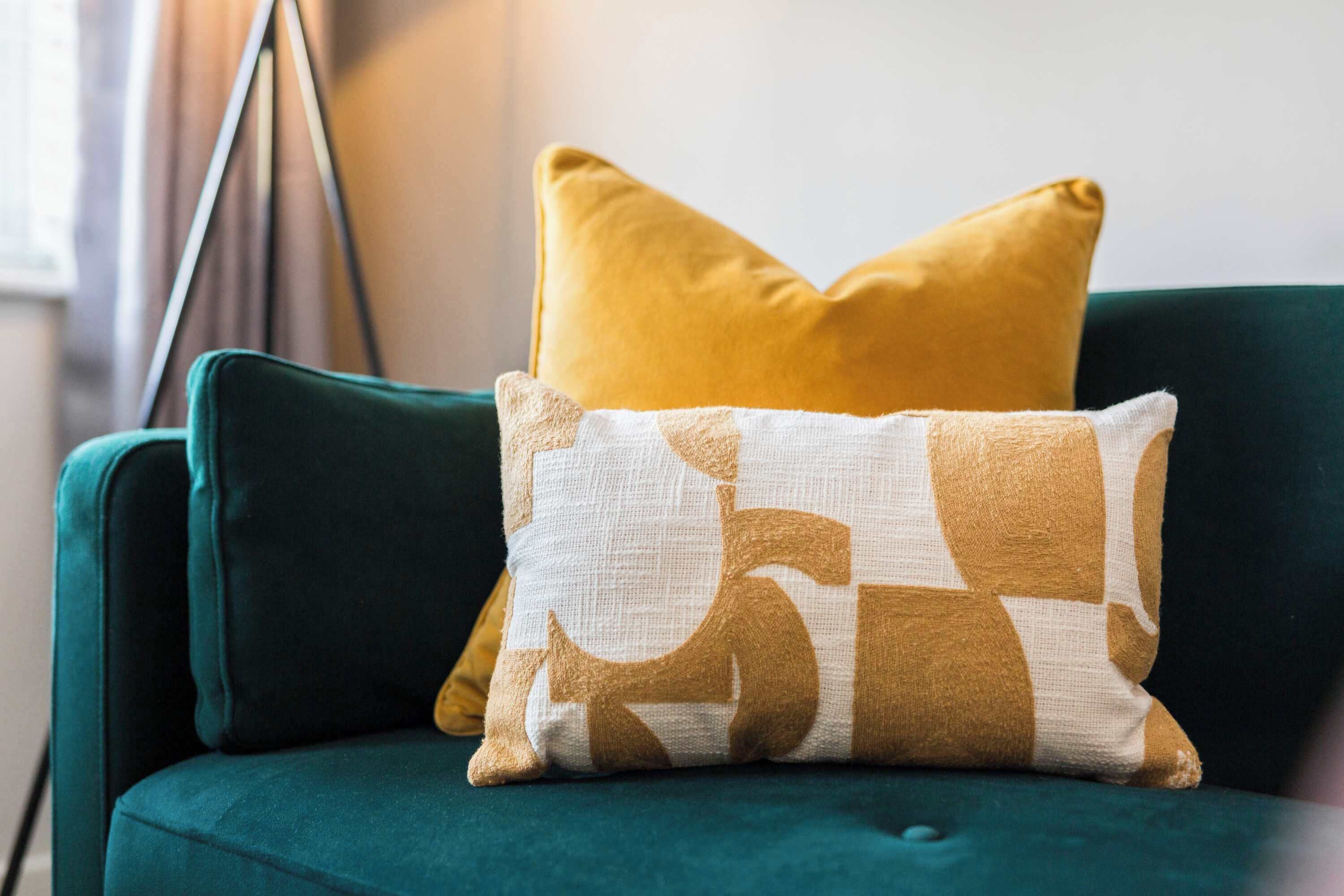 A teal sofa with two pillows, one yellow and one patterned with abstract shapes.