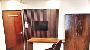 A room with a wooden desk, a black chair, a TV mounted on a wooden panel wall, and a wardrobe. - Hotel the terminal (New Delhi)