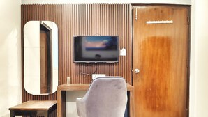 A room with a wooden door, a television mounted on the wall, a grey chair, a small wooden table, and a mirror. - Hotel the terminal (New Delhi)