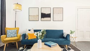 Un salon avec un fauteuil jaune, un canapé bleu, une table basse ornée d’un vase et de livres, et trois œuvres d’art encadrées accrochées au mur.