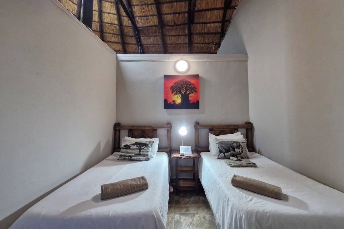 A small, cozy room with two single beds, a wooden headboard, a welcome sign, and a painting of a tree.