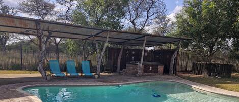 A swimming pool with a brick border, surrounded by a covered patio area with blue chairs.