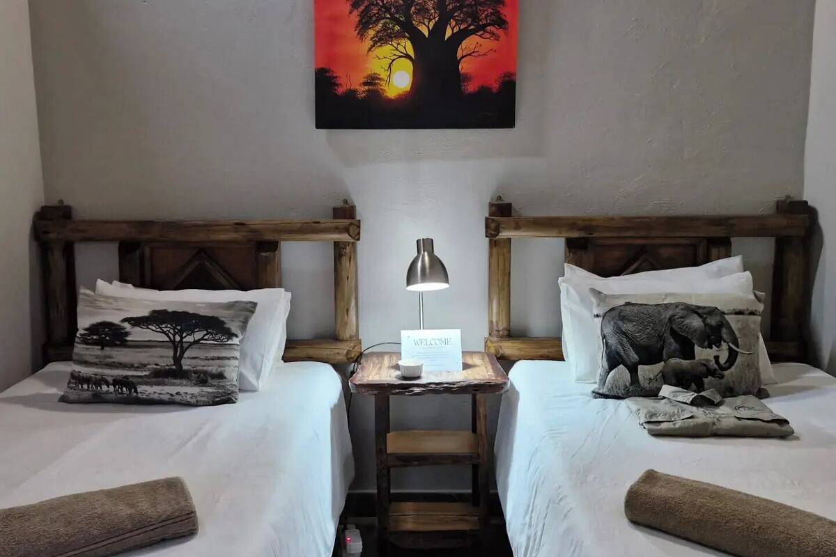 A room with two beds, each with a unique pillow design, a small table with a lamp, and a painting of a tree at sunset on the wall.