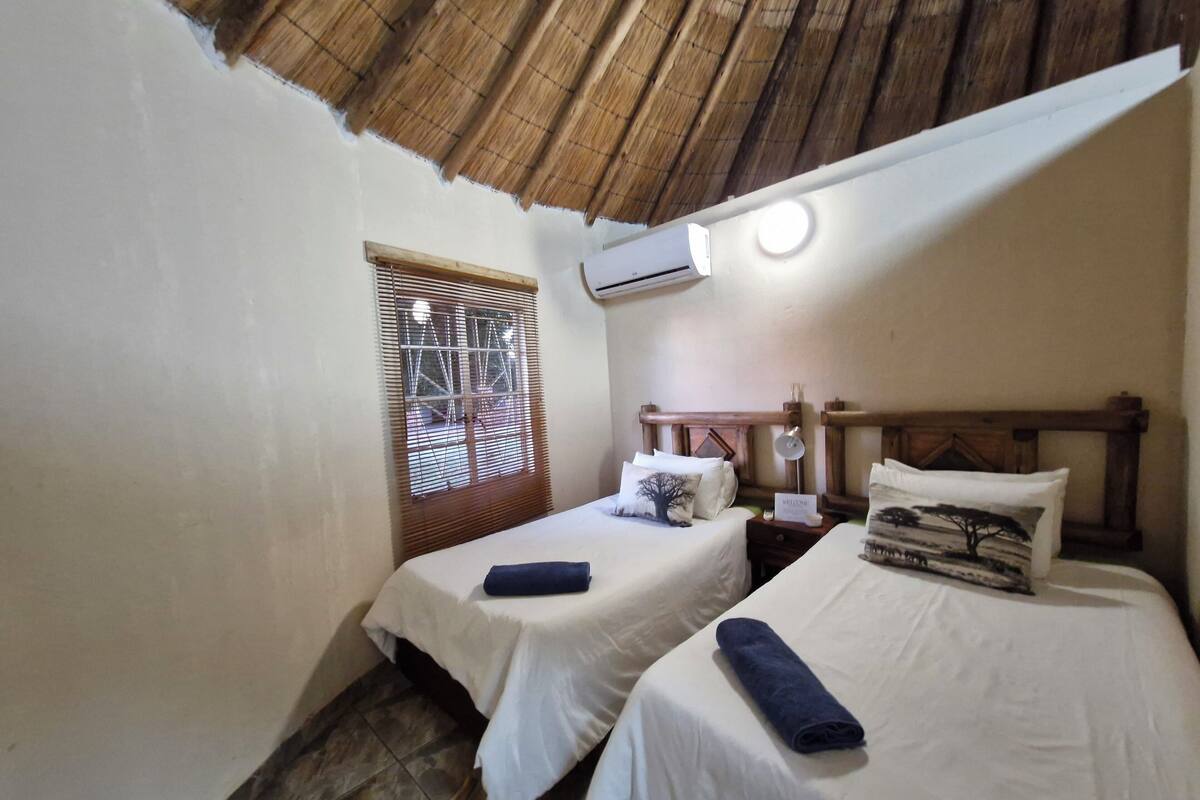 A small, cozy room with two single beds, a wooden headboard, a window with bamboo blinds, and a ceiling fan.