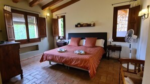 Un dormitorio con cama, cabecera de madera, mesitas de noche, una silla, un escritorio y ventana con cortinas.