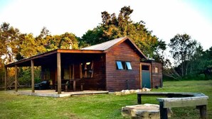 A rustic wooden cabin with a covered porch, a stone bench, and a small table in a grassy area. - Enchanting Forest Cabin in Tsitsikamma (Stormsriver, Tsitsikamma)