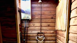A small, rustic bathroom with wooden walls, a white toilet, a heart-shaped decoration, and a stone pathway leading to a shower area. - Enchanting Forest Cabin in Tsitsikamma (Stormsriver, Tsitsikamma)