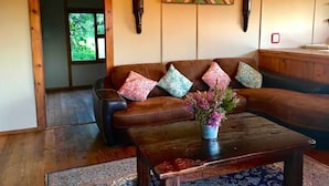 A living room with a brown leather sofa, colorful cushions, a wooden coffee table with a flower vase, and a geometric wall art piece. - Enchanting Forest Cabin in Tsitsikamma (Stormsriver, Tsitsikamma)