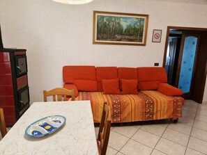 A living room with a sofa, a dining table, and a chair. There is a painting on the wall and a door with a frosted glass panel.