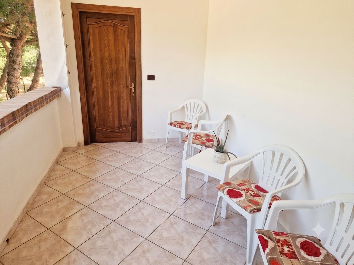 Un balcón con puerta de madera, sillas de plástico blanco y piso de baldosas.