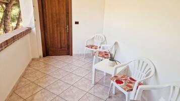 Un balcón con puerta de madera, sillas de plástico blanco y piso de baldosas.